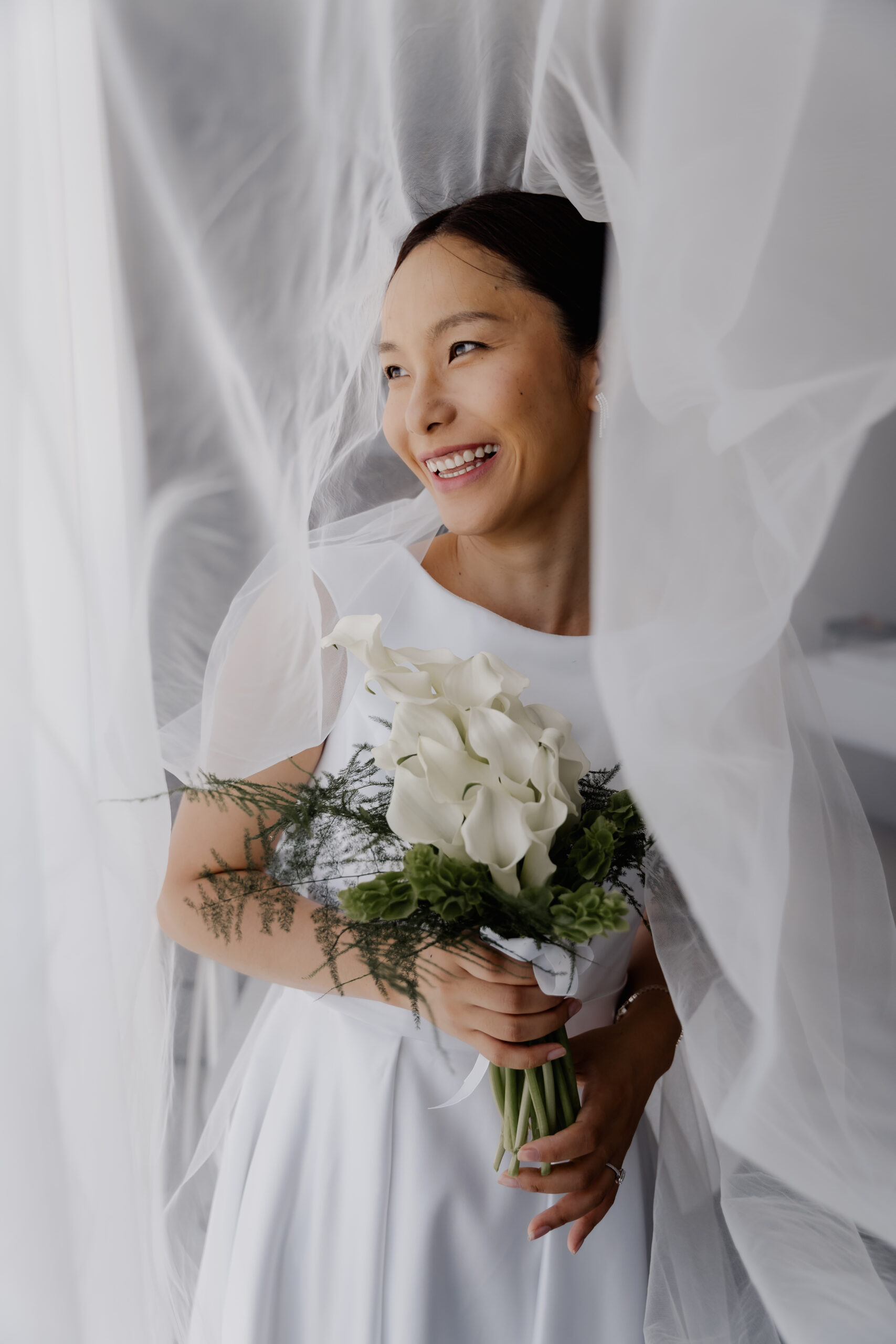 Eine Braut in einem weißen Kleid, die Calla-Lilien in den Händen hält, lächelt und deren transparenter Schleier um sie herum weht – perfekt in Szene gesetzt von ihrem aufmerksamen Hochzeitsplaner.