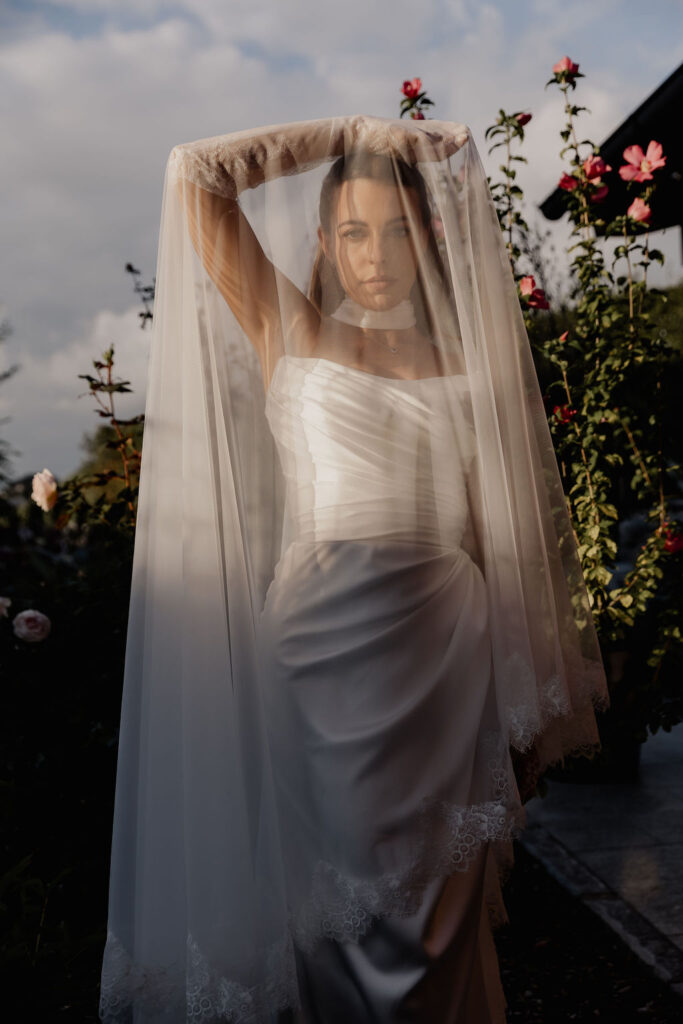 Eine Braut in einem weißen Brautkleid steht im Freien bei ihrer Hochzeit im Ausland und hält sich inmitten blühender Blumen einen durchsichtigen Schleier vor das Gesicht.