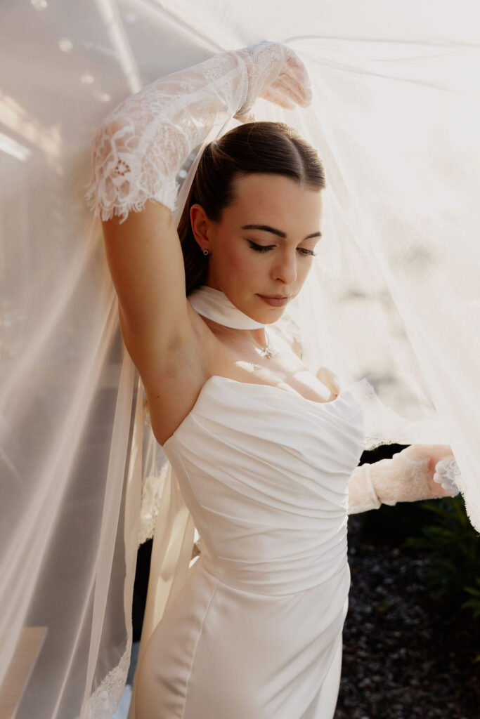 Eine Braut in einem weißen Brautkleid und Spitzenhandschuhen posiert an der frischen Luft anmutig unter einem wallenden Schleier und verkörpert damit die Romantik einer Hochzeit im Ausland.