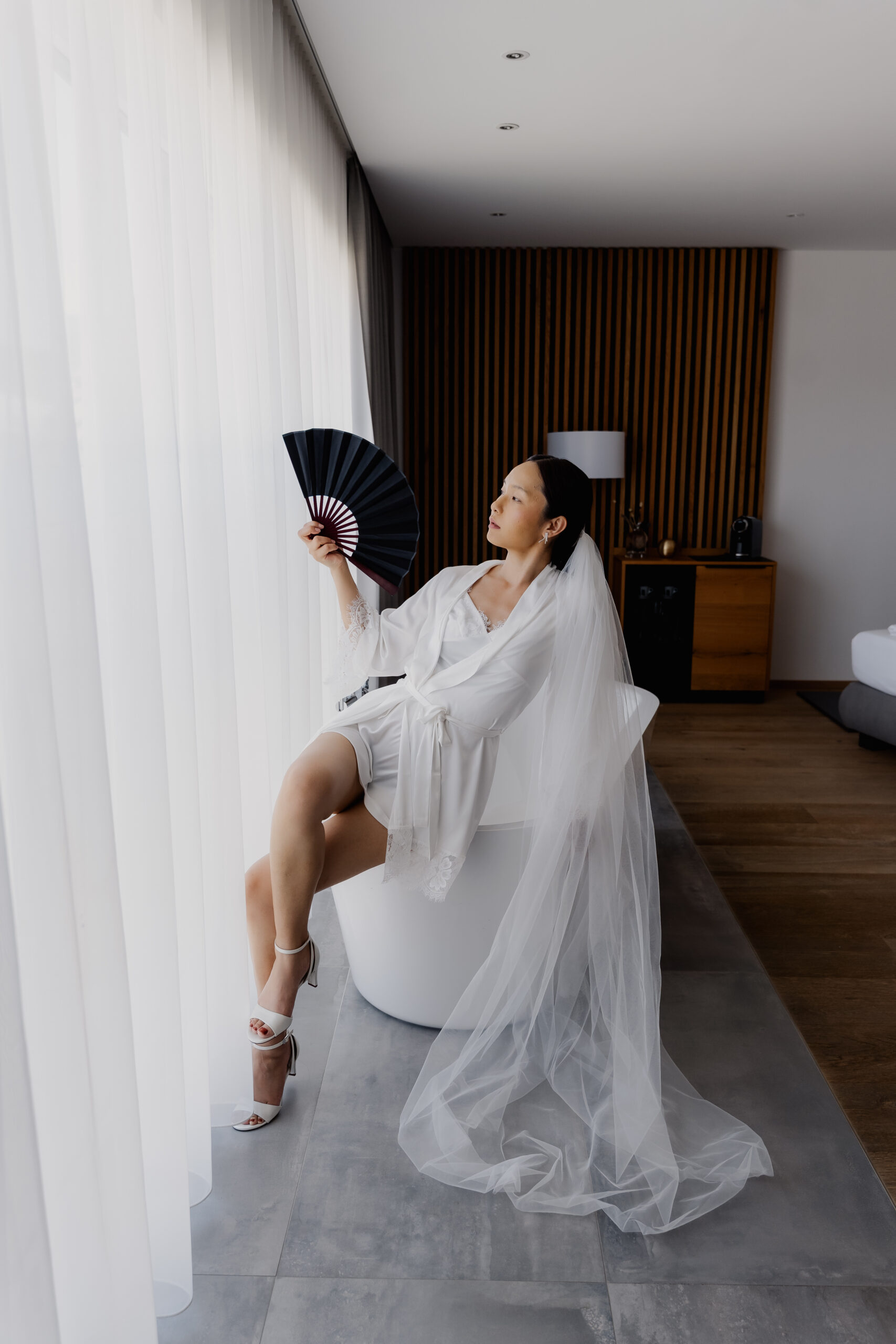 Eine Braut in einem Brautkleid und mit Schleier sitzt in einer Badewanne, hält einen schwarzen Fächer in der Hand und blickt aus dem Fenster eines modernen Zimmers, während sie nachdenklich den kommenden Momenten entgegenfiebert und ihre Hochzeitsplanerin sich um jedes Detail kümmert.