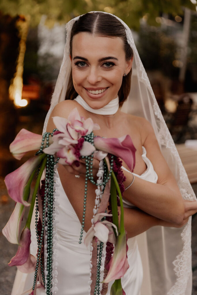 Eine lächelnde Braut in einem weißen Kleid, die einen rosa Blumenstrauß in der Hand hält und einen Spitzenschleier trägt, im Freien bei ihrer unvergesslichen Hochzeit an einem besonderen Ort.