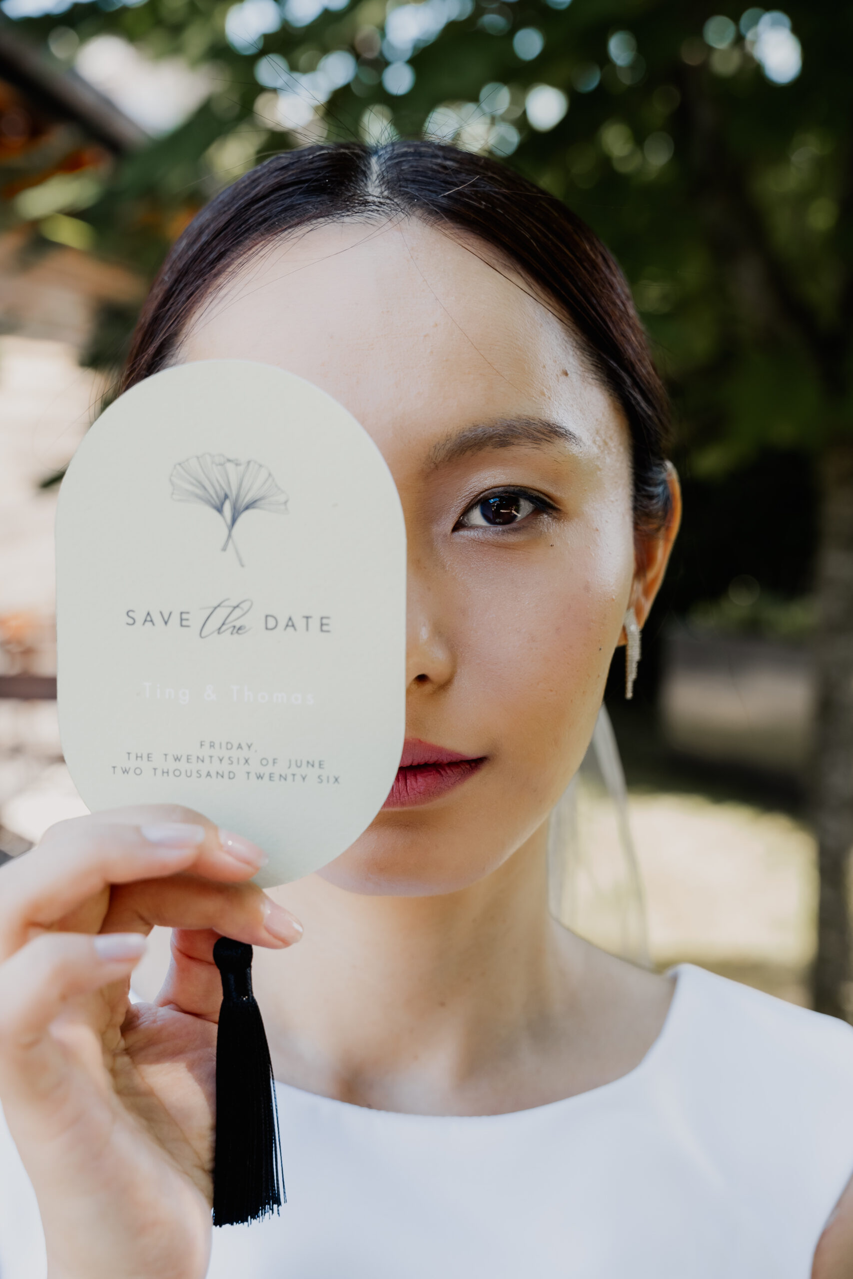 Eine Frau in Weiß, vielleicht eine Hochzeitsplanerin oder Eventmanagerin, hält eine „Save the Date“-Karte in der Hand, die ihr Gesicht zur Hälfte verdeckt, während sie im Freien steht, hinter ihr ist Grün zu sehen.