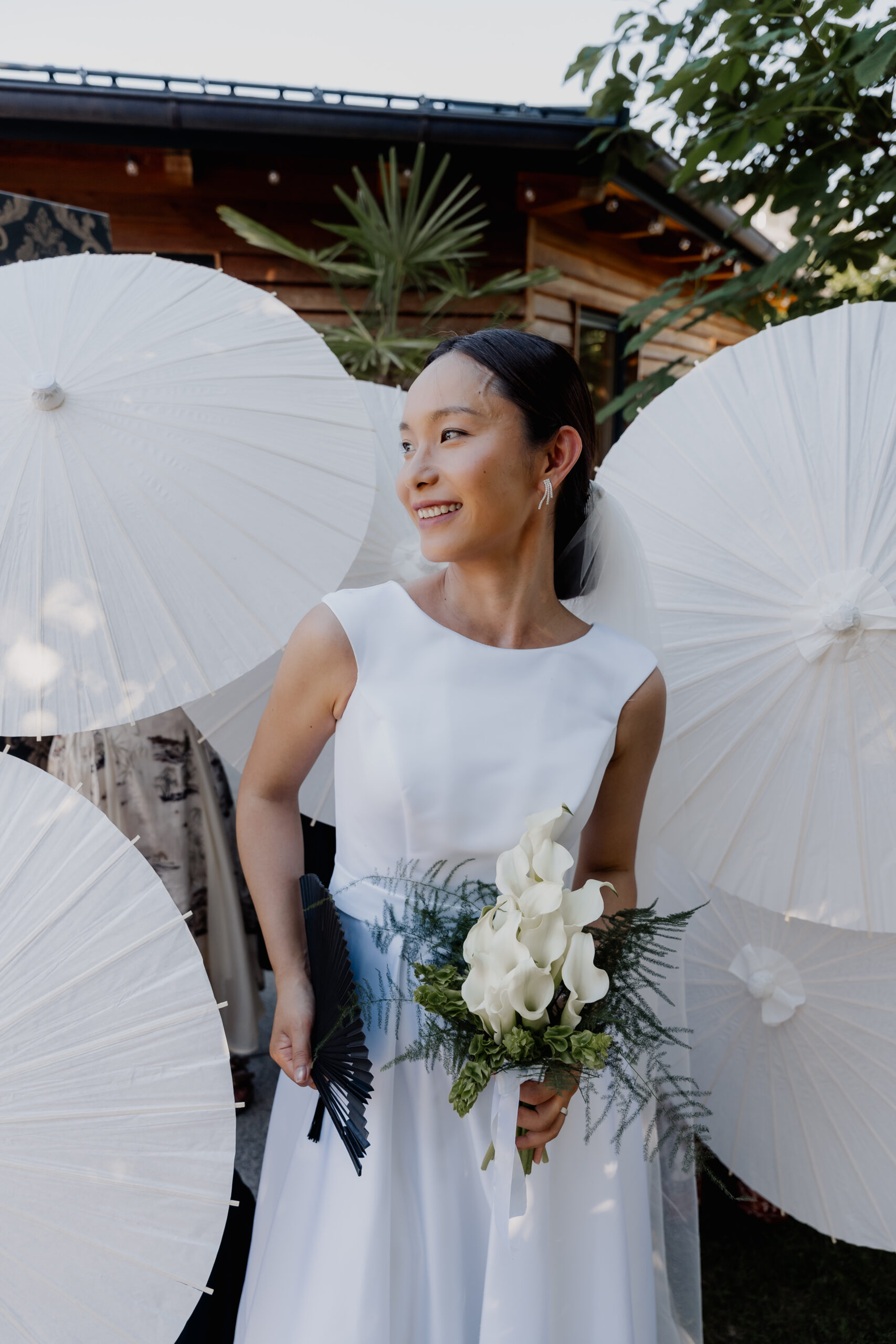 Eine Braut in einem weißen Kleid, die Calla-Lilien in den Händen hält, steht im Freien vor weißen Sonnenschirmen und lächelt – ihr freudiger Moment wurde von einem erfahrenen Hochzeitsplaner perfekt inszeniert.