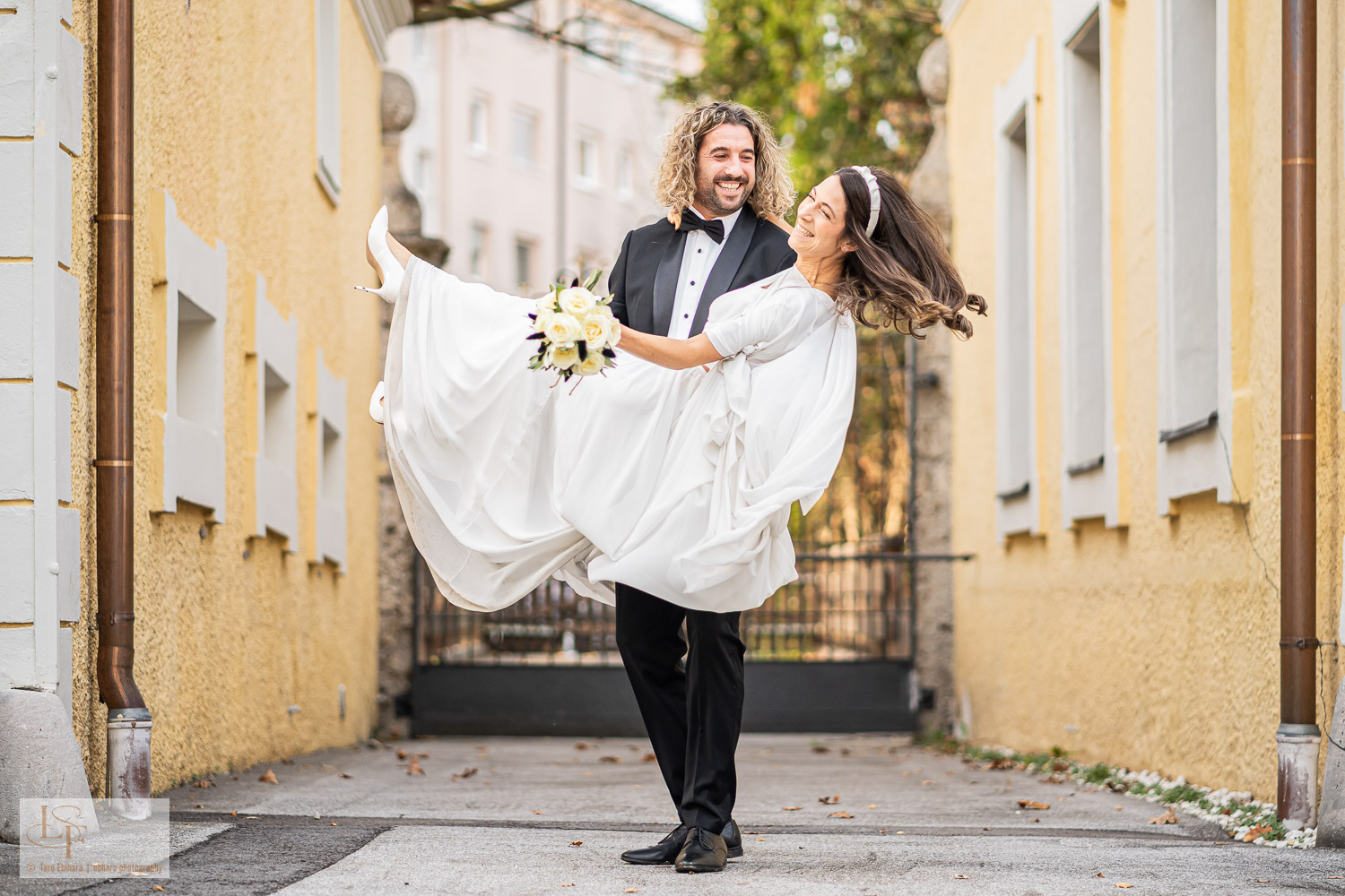 Ein Bräutigam im Smoking hebt eine lächelnde Braut im weißen Kleid hoch, während die beiden fröhlich im Freien posieren und so den Zauber ihres besonderen Tages an einer der atemberaubenden Hochzeitslocations in Salzburg Stadt festhalten.