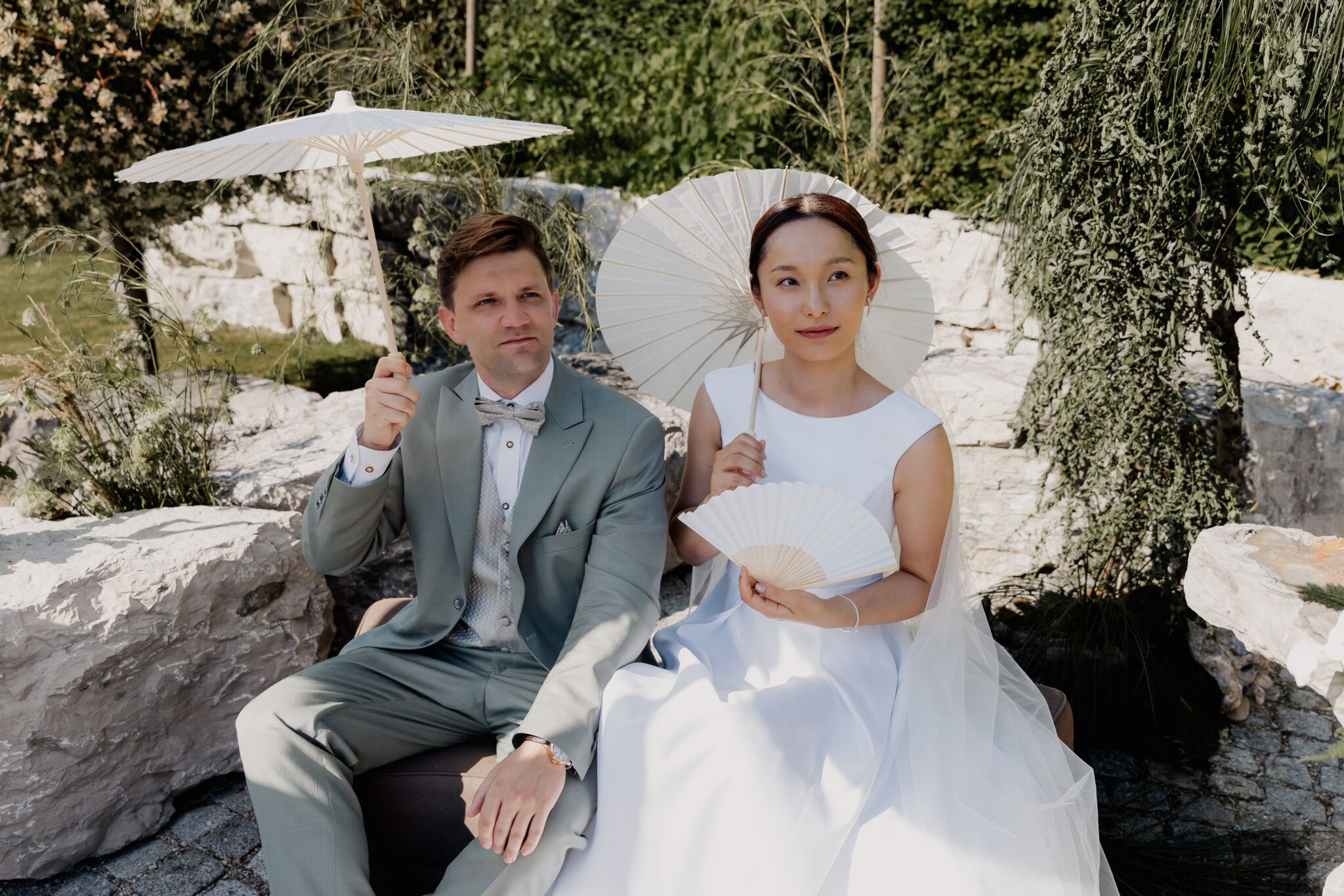 Braut und Bräutigam sitzen im Freien mit Sonnenschirmen und Fächern, in festlicher Hochzeitskleidung, umgeben von Felsen – eine malerische Szene, die von einem erfahrenen Hochzeitsplaner sorgfältig inszeniert wurde.