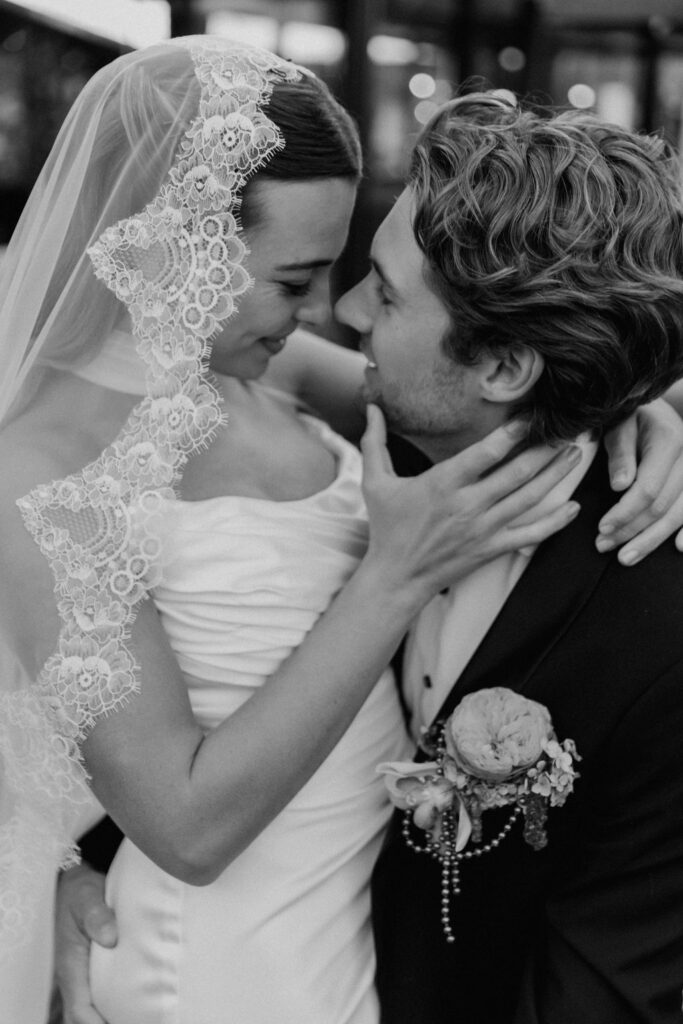 Ein Brautpaar umarmt sich innig und lächelt sich liebevoll an; die Braut trägt bei ihrer Hochzeit am Reiseziel einen Spitzenschleier.