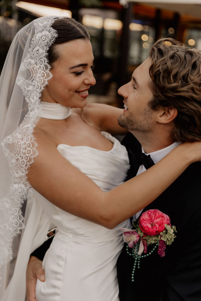 Die Braut in weißem Kleid und Schleier umarmt den Bräutigam im schwarzen Anzug während ihrer Hochzeit im Ausland; beide lächeln und blicken sich liebevoll an.
