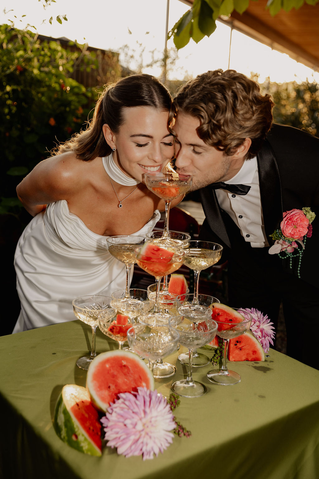Braut und Bräutigam trinken aus dem obersten Champagnerglas eines Turms, während Wassermelonenscheiben und Blumen den Tisch schmücken – ein Bild, das die romantische Atmosphäre einer Hochzeit im Ausland perfekt einfängt.