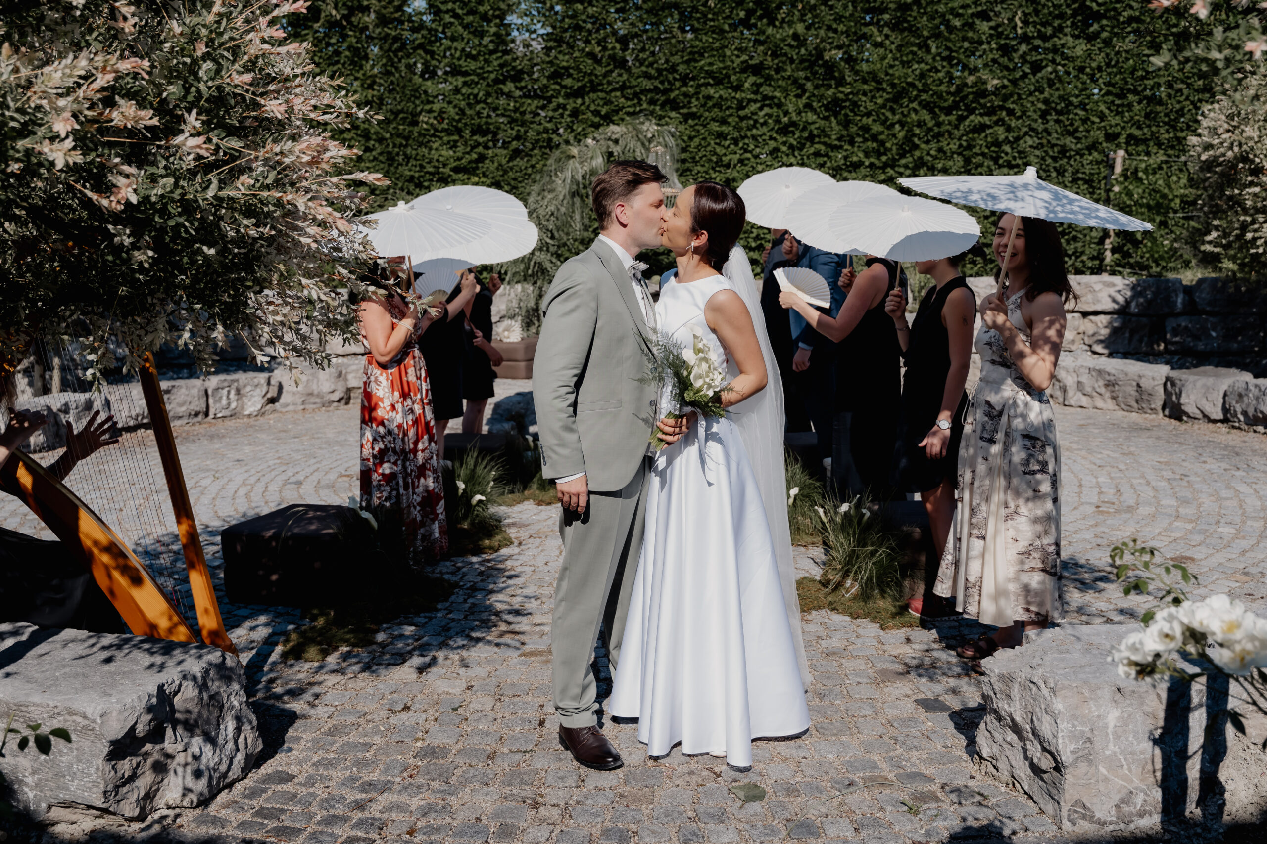 Ein Brautpaar küsst sich im Freien, während im Hintergrund die Gäste weiße Sonnenschirme halten – ein idyllischer Moment, der von einem professionellen Hochzeitsplaner perfekt inszeniert wurde.