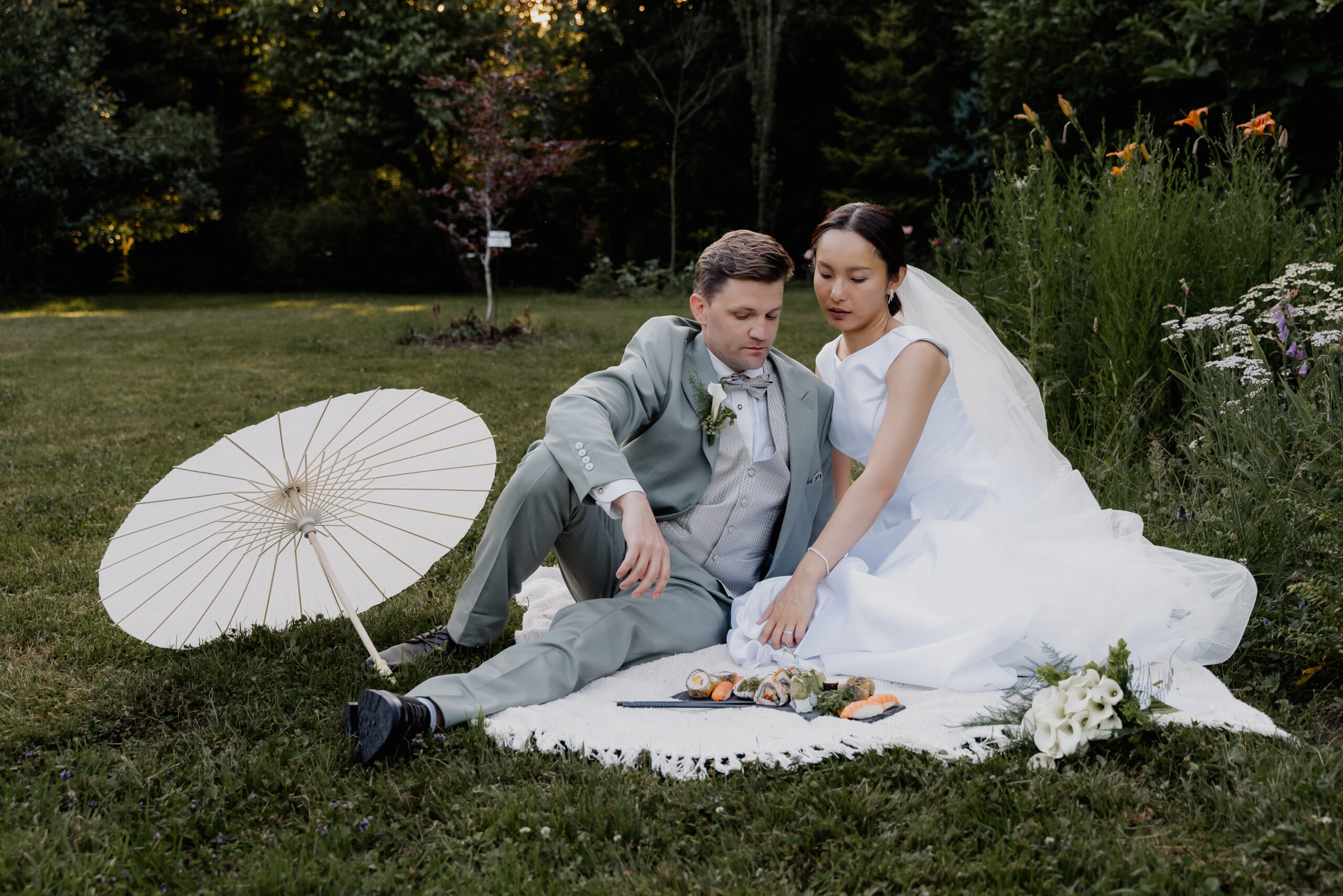 Braut und Bräutigam sitzen draußen auf einer Picknickdecke, neben ihnen liegen Sushi, der Brautstrauß und ein weißer Sonnenschirm – ihr ganz privater Moment wurde von einem erfahrenen Hochzeitsplaner sorgfältig inszeniert.