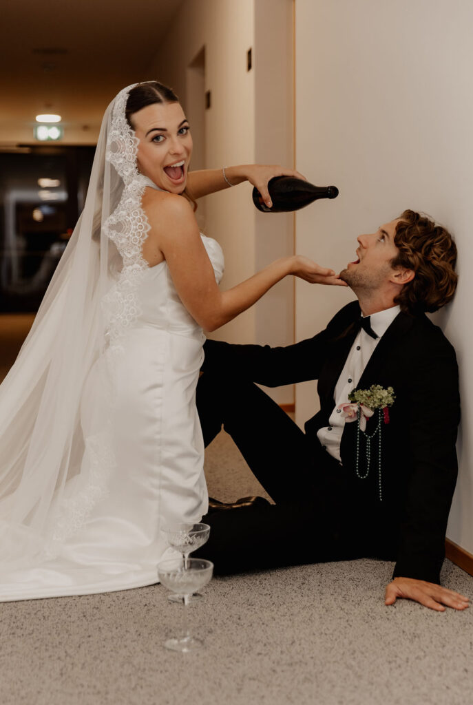 Die Braut gießt dem Bräutigam spielerisch Champagner in den Mund – in einem Flur, beide lachend und in Hochzeitskleidung – und fängt damit die fröhliche Stimmung dieser Hochzeit in Salzburg ein, die von Stephanie Weddings meisterhaft dokumentiert wurde.
