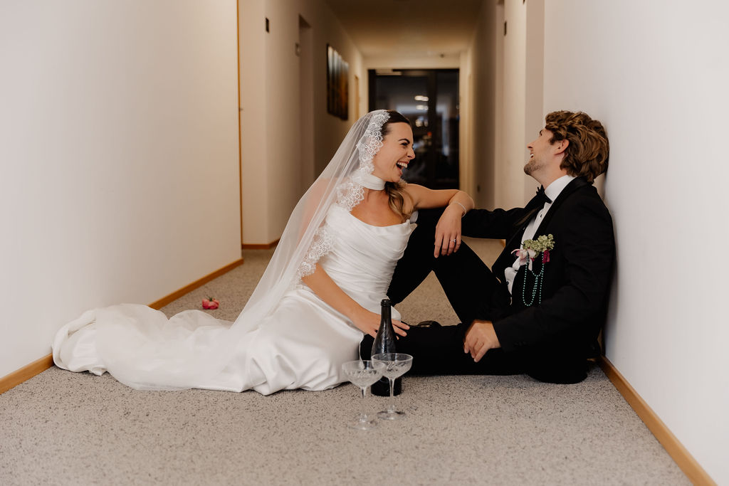 Braut und Bräutigam sitzen auf dem Boden in einem Flur, lachen gemeinsam, vor sich ihre Champagnergläser, und genießen die Freude und Spontaneität ihrer Hochzeit am Reiseziel.