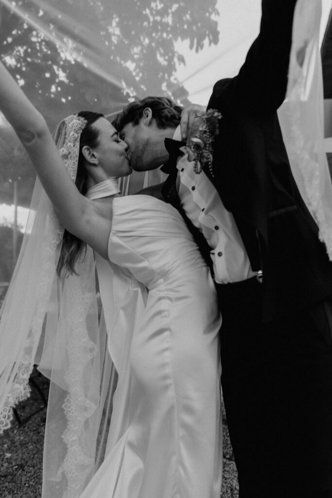 Ein Brautpaar tauscht an seinem Hochzeitstag im Ausland unter einem Schleier einen freudigen Kuss aus, die Arme feierlich in die Höhe gestreckt.