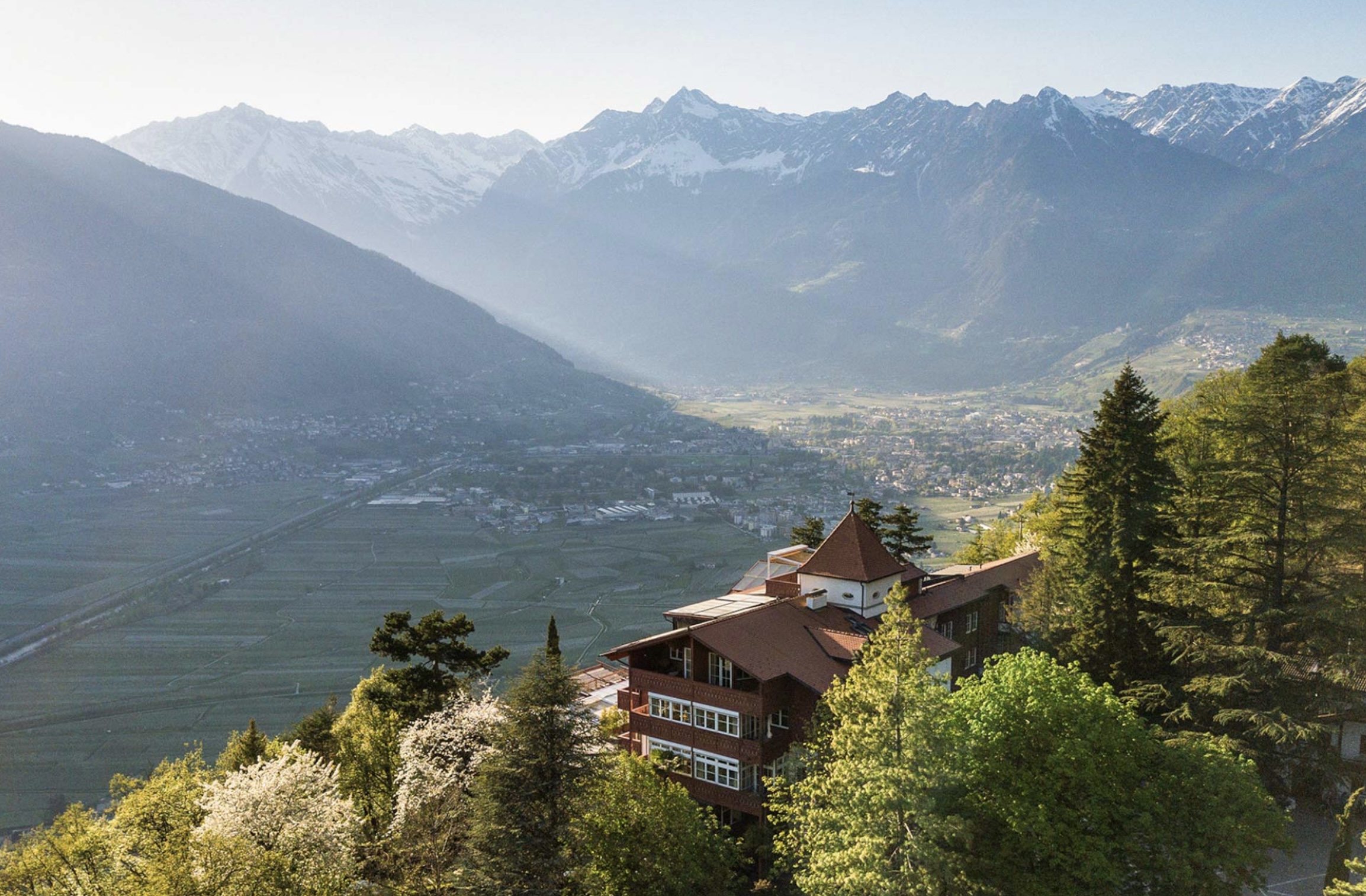 Ein großes Holzhaus auf einem Hügel überragt unter strahlend blauem Himmel ein Tal mit Bergen im Hintergrund und ist damit eine der reizvollsten Hochzeitslocations in Südtirol.