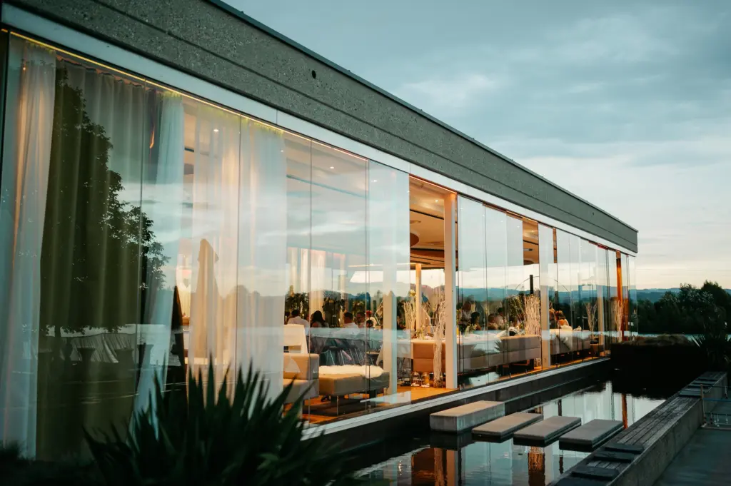 Ein modernes Restaurant mit Glasfassade, in dem sich bei Sonnenuntergang das Licht im Wasser spiegelt, während drinnen Gäste speisen und draußen üppiges Grün zu sehen ist – eine ideale Hochzeitslocation in Oberösterreich für eine unvergessliche Hochzeit oder elegante Feierlichkeiten an einem der atemberaubendsten Hochzeitsorte der Region.