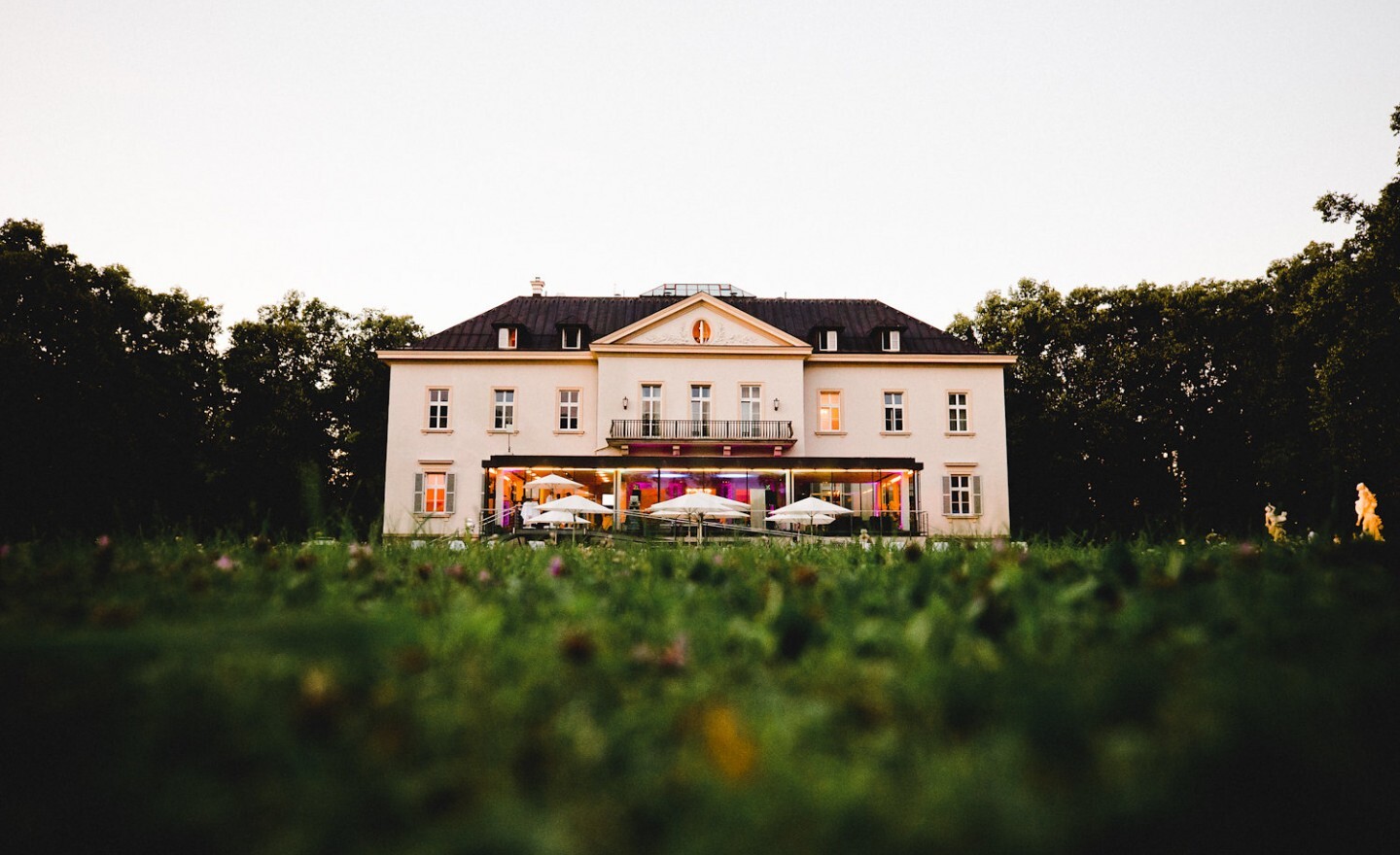 Ein großes weißes Herrenhaus mit Balkon, beleuchteten Fenstern und Sonnenschirmen – einer der bezauberndsten Hochzeitsorte in Salzburg Stadt –, umgeben von Rasenflächen und Bäumen.