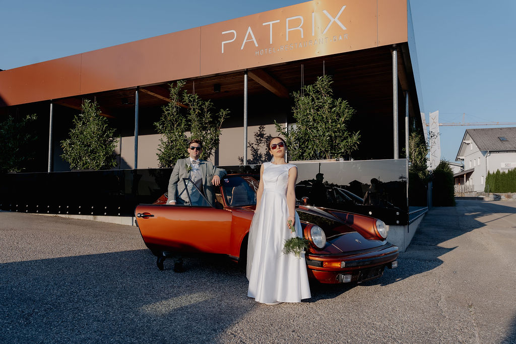 Ein Brautpaar posiert neben einem roten Oldtimer vor dem Patrix Hotel, einem der reizvollen Orte für eine Hochzeit an einem sonnigen Tag.