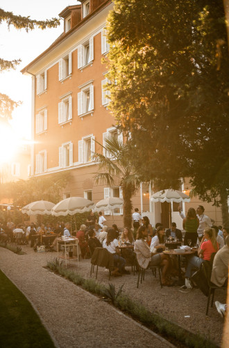 Menschen, die in der Abendsonne vor einem Restaurant im Freien speisen, in der Nähe eines hohen orangefarbenen Gebäudes mit weißen Fenstern, das an die bezaubernden Hochzeitslocations in Südtirol erinnert.