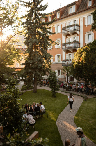 Die Gäste speisen und spazieren in einem sonnendurchfluteten Gartenhof neben einem hohen, orangefarbenen Gebäude mit Balkonen und Bäumen – eine einladende Kulisse, die den Charme der Hochzeitslocations in Südtirol einfängt.