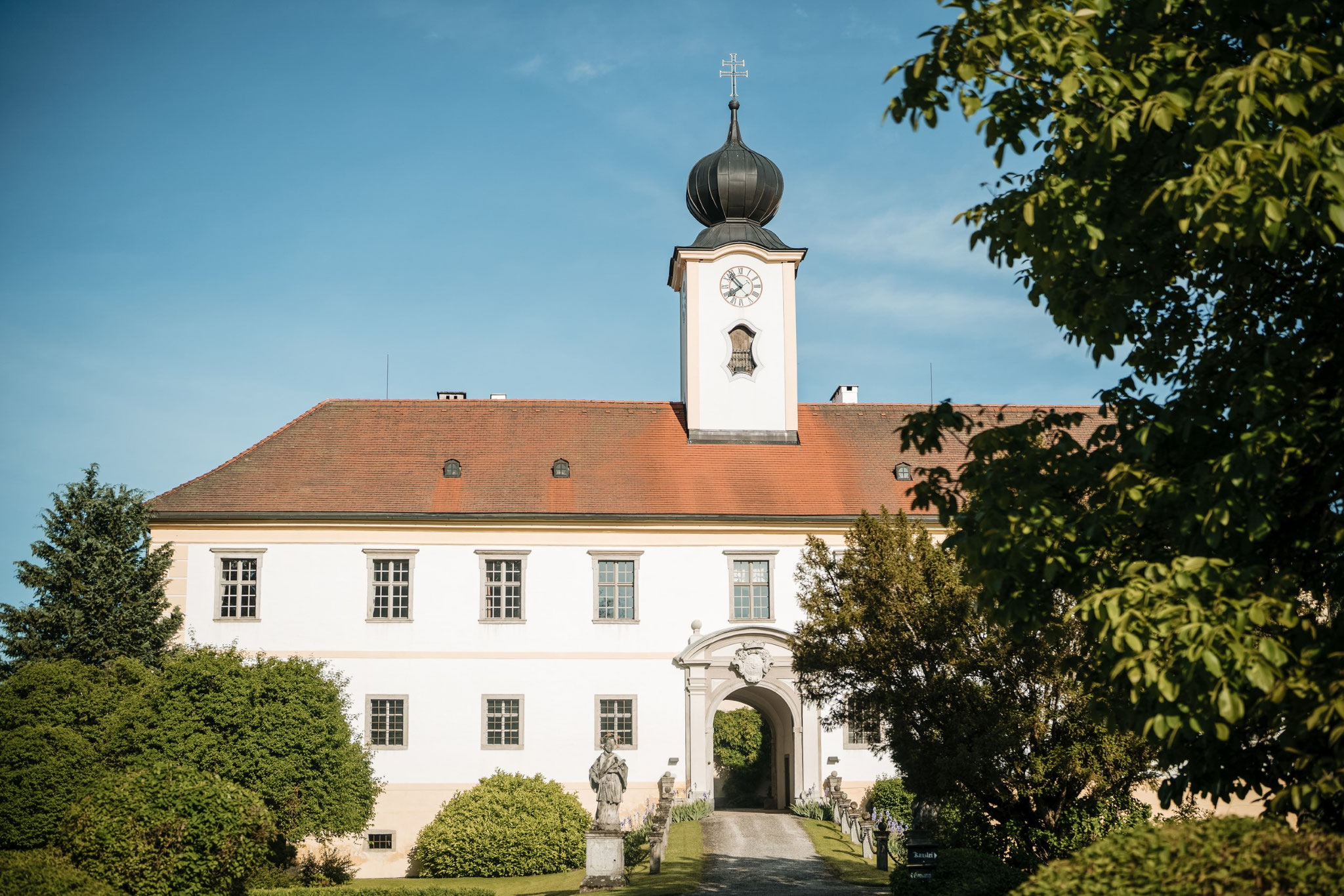 Ein weißes Gebäude mit rotem Dach und Uhrturm, umgeben von Bäumen und viel Grün unter blauem Himmel – einer der schönsten Orte für Ihre Hochzeit und eine begehrte Hochzeitslocation in Oberösterreich.
