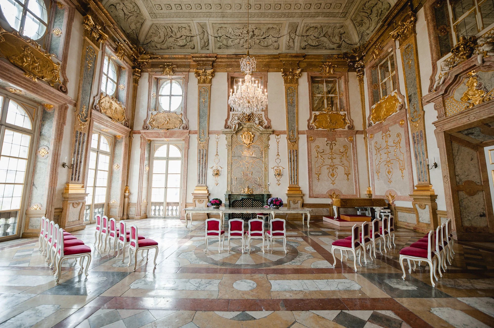 Ein prunkvoller Ballsaal mit Kronleuchter, goldenen Akzenten, hohen Fenstern und Reihen rot gepolsterter Stühle, die im Kreis angeordnet sind – eine der elegantesten Hochzeitslocations in Salzburg Stadt.