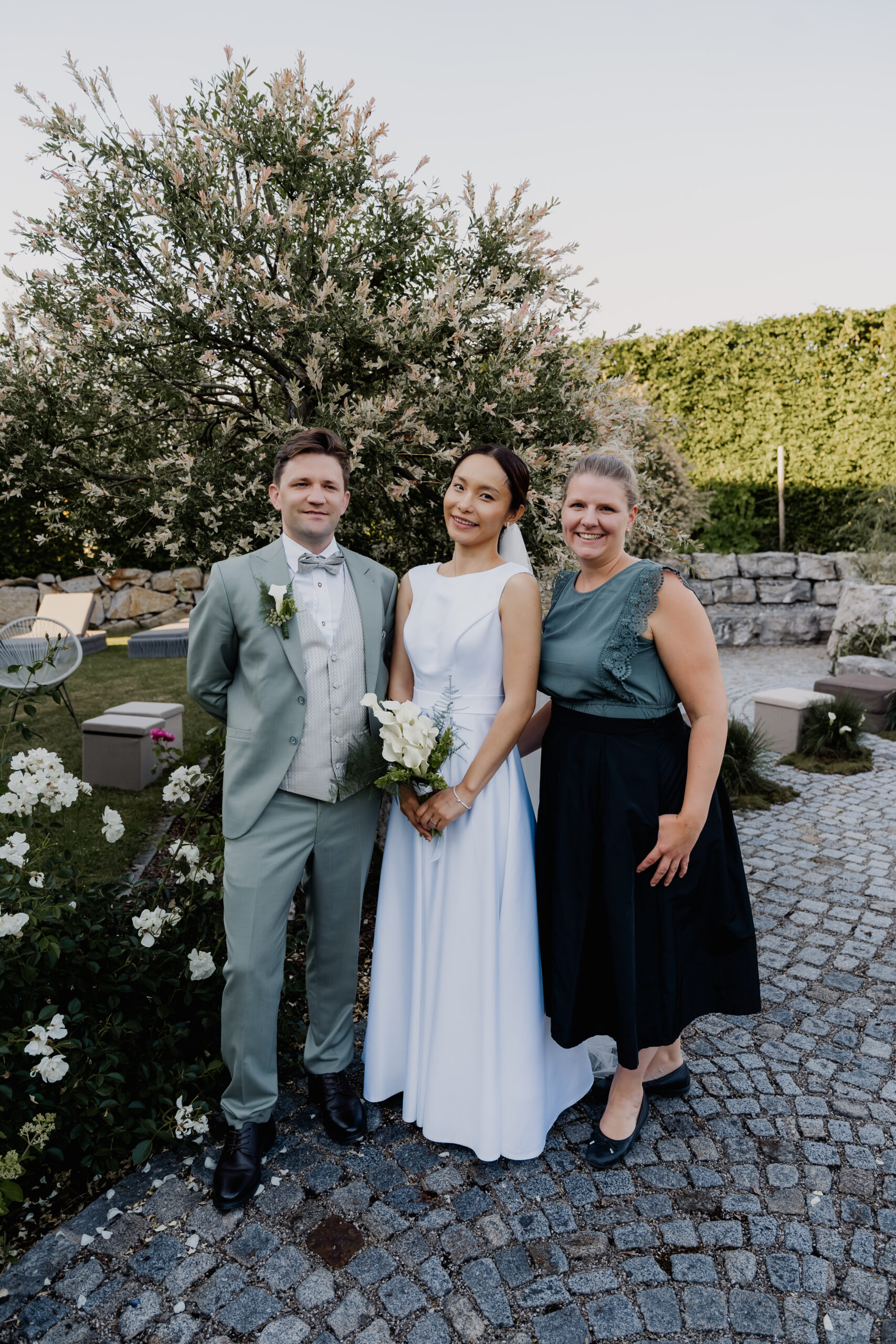 Die Braut im weißen Kleid, der Bräutigam im grauen Anzug und eine Frau in einem grünen Oberteil – möglicherweise die Hochzeitsplanerin – posieren draußen auf einem Steinweg.
