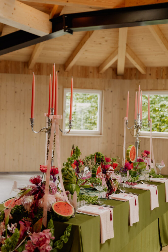 Ein langer Tisch mit grünem Tischtuch, rosa Kerzen, Blumen und Wassermelonenscheiben sorgt in einem holzgetäfelten Raum für eine bezaubernde Atmosphäre – die perfekte Inspiration für Hochzeitslocations in Salzburg Stadt.